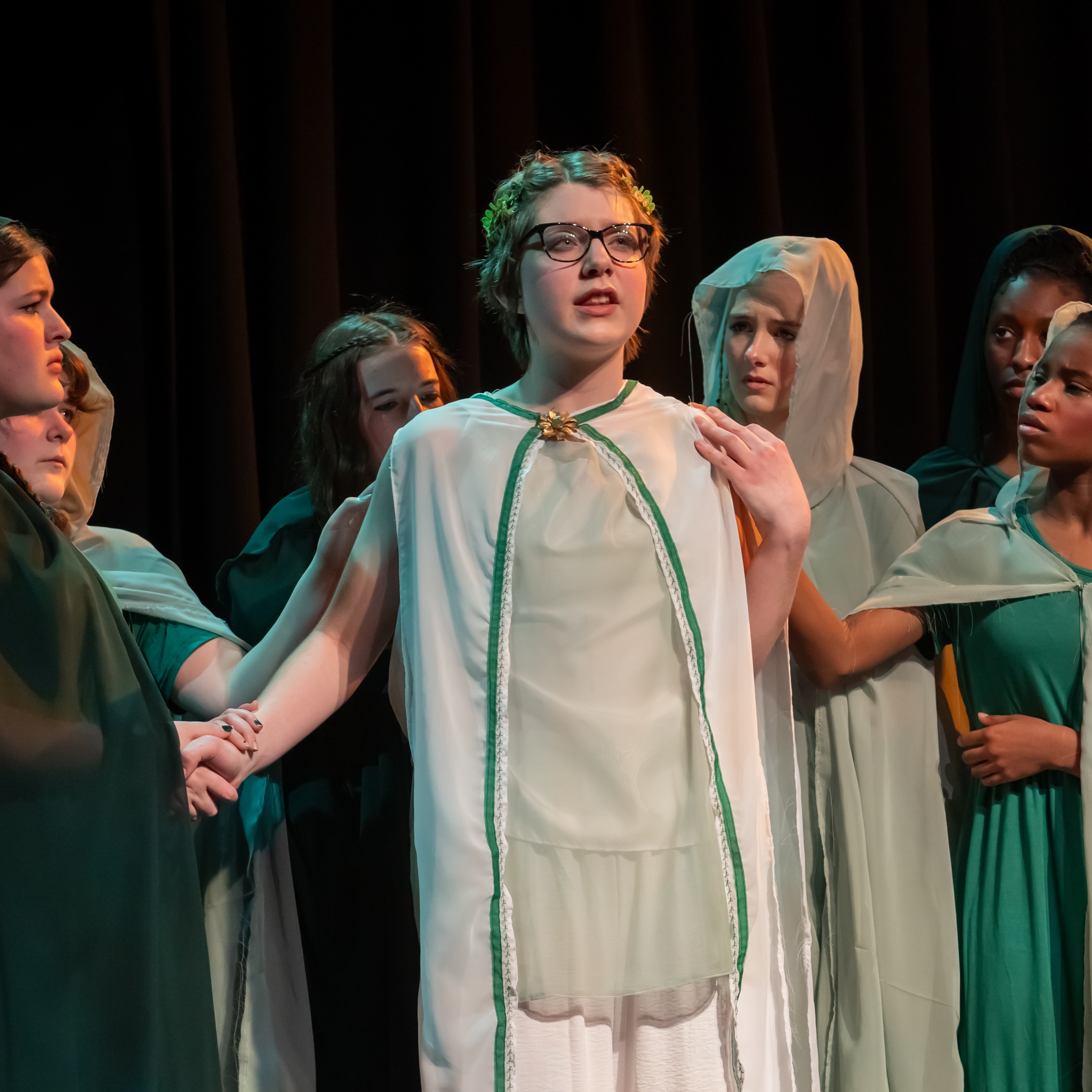 A young woman in a white robe stares at our upper right. She is surrounded by oither women in green robes who look at her.